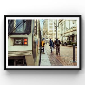 A transit train car passes pedestrians walking along a narrow urban street with tall buildings and storefronts.