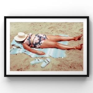 A vintage photograph shows a person in a floral swimsuit relaxing on a beach towel with shoes and a hat nearby.