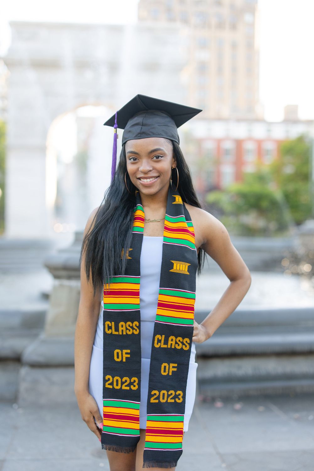 Graduate in cap and gown with kente cloth stole celebrating near Washington Square Park.
