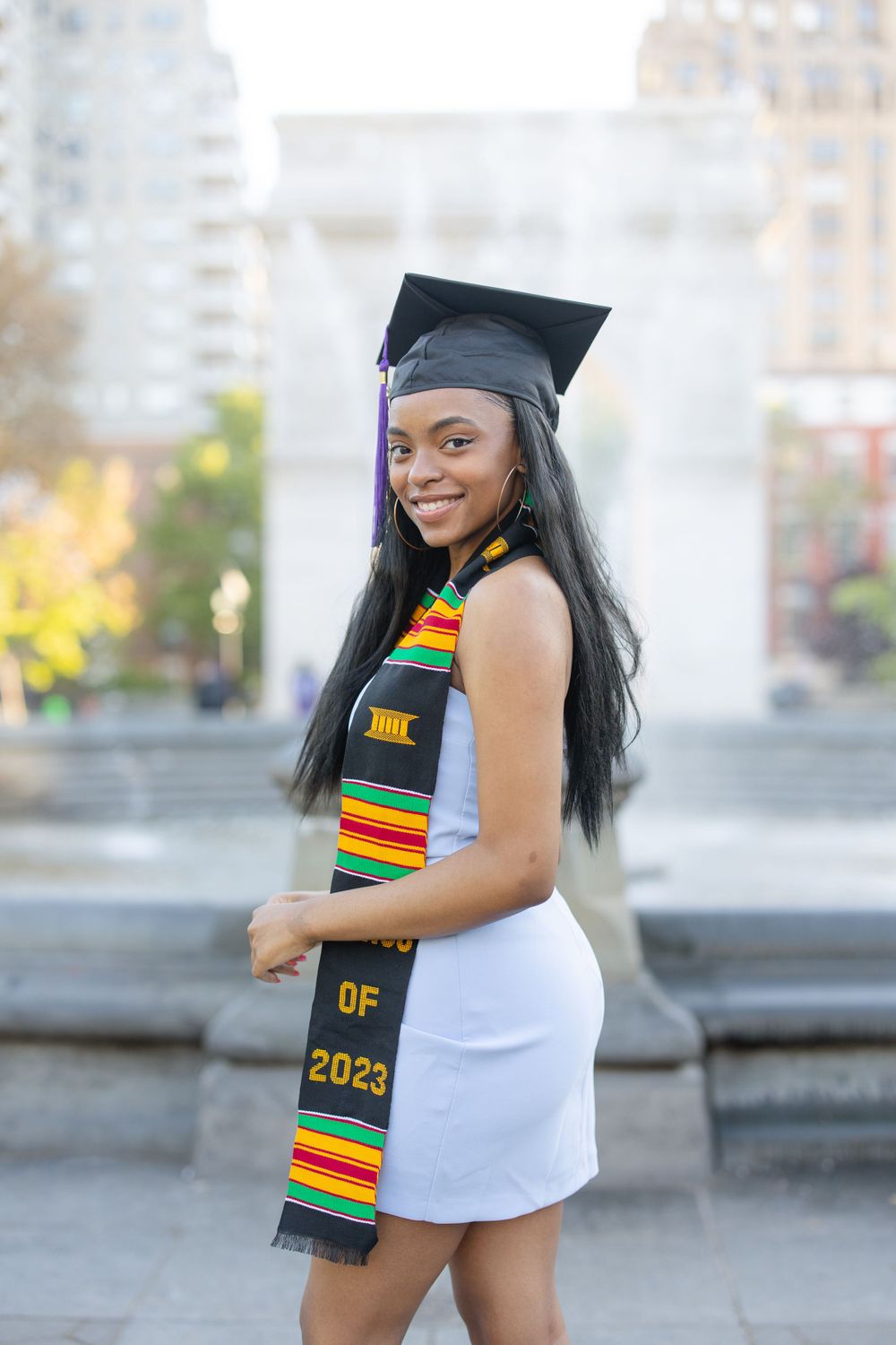 Graduate in white dress and kente stole smiling in front of Washington Square Arch.