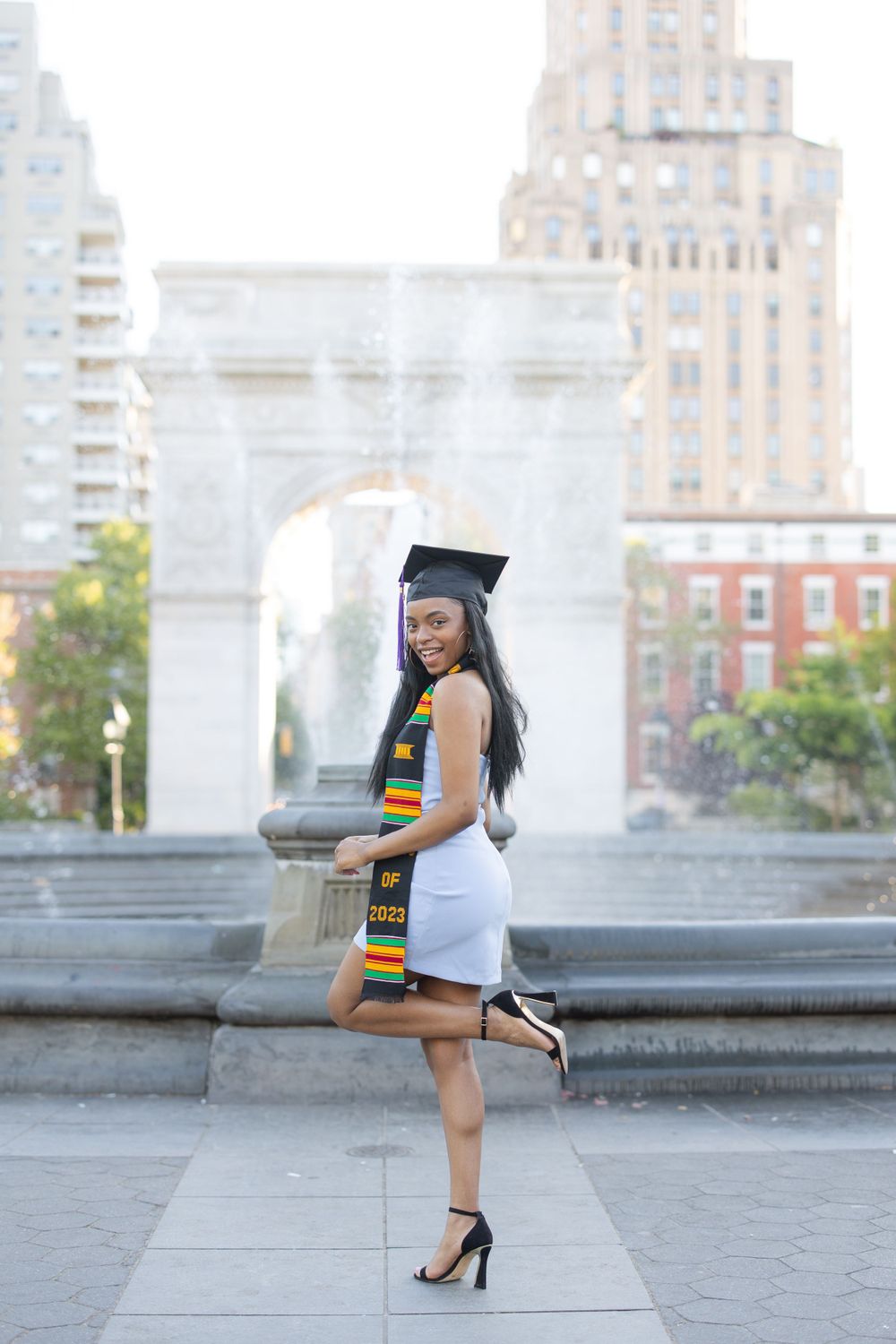 Graduate striking a pose in cap and gown near Washington Square Park fountain.