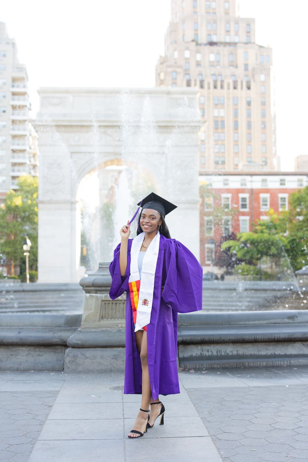 Graduate in purple graduation robe posing by Washington Square Park fountain.