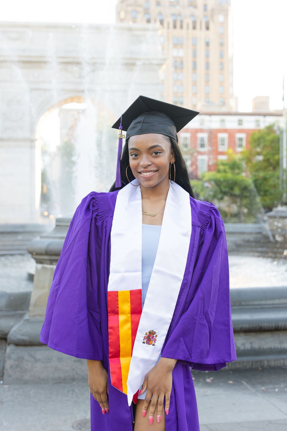 Graduate in purple regalia and kente stole celebrating near a fountain.