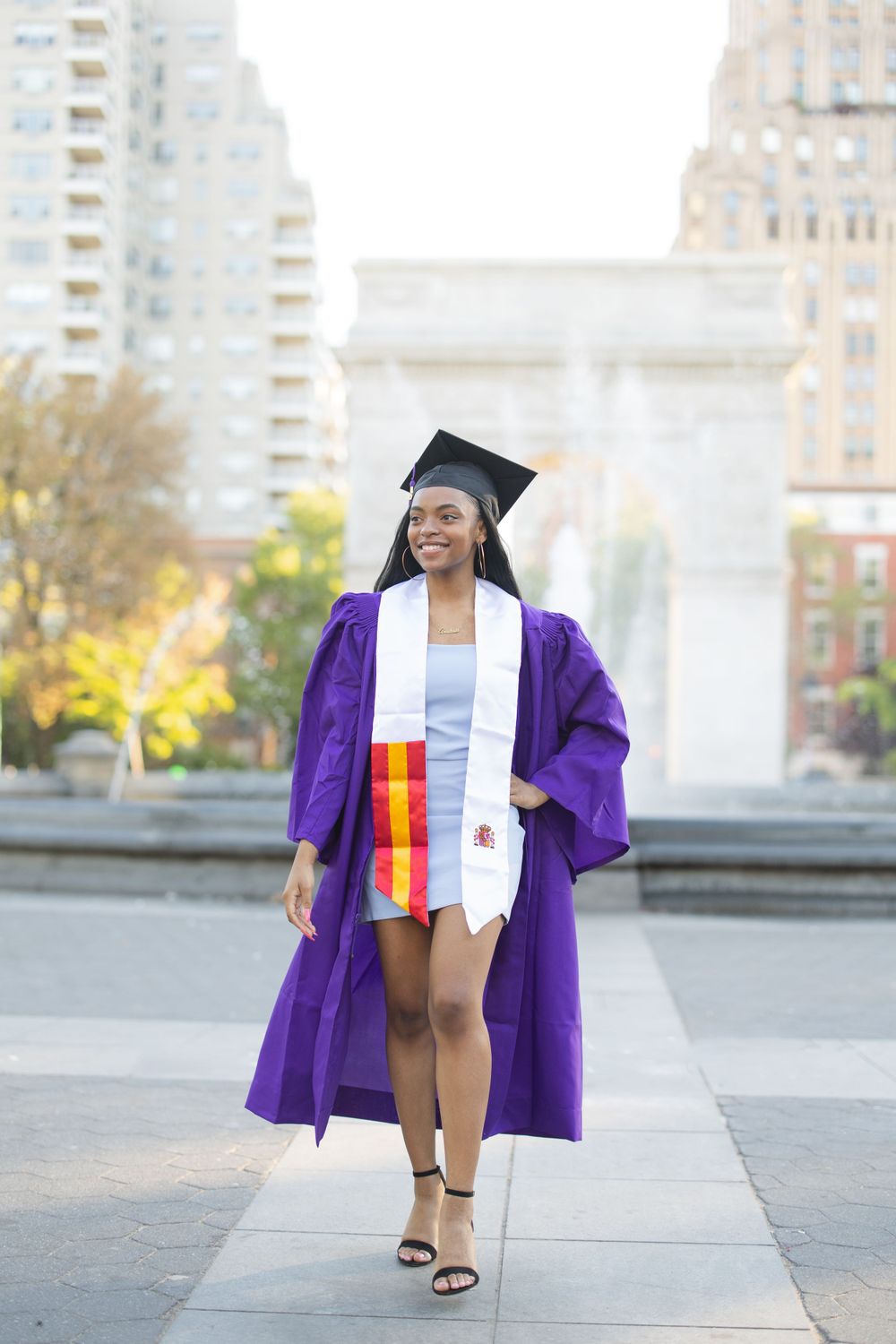 Graduate wearing purple commencement robe and graduation cap poses in front of Washington Square Arch.