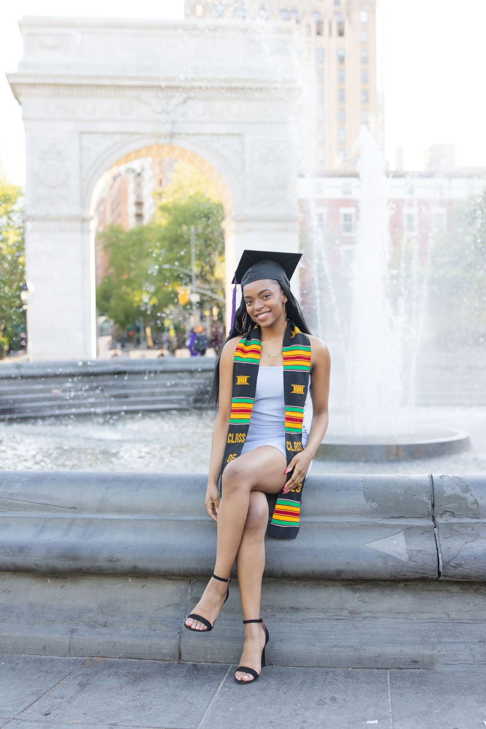 Graduate in traditional regalia with colorful stole sits by fountain at Washington Square Park.