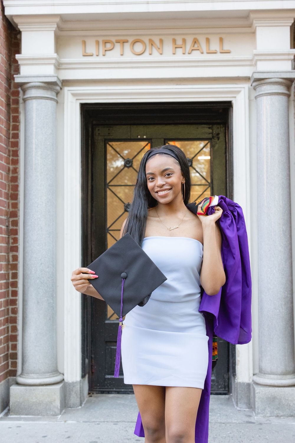 Graduate poses with diploma and gown outside Lipton Hall entrance.
