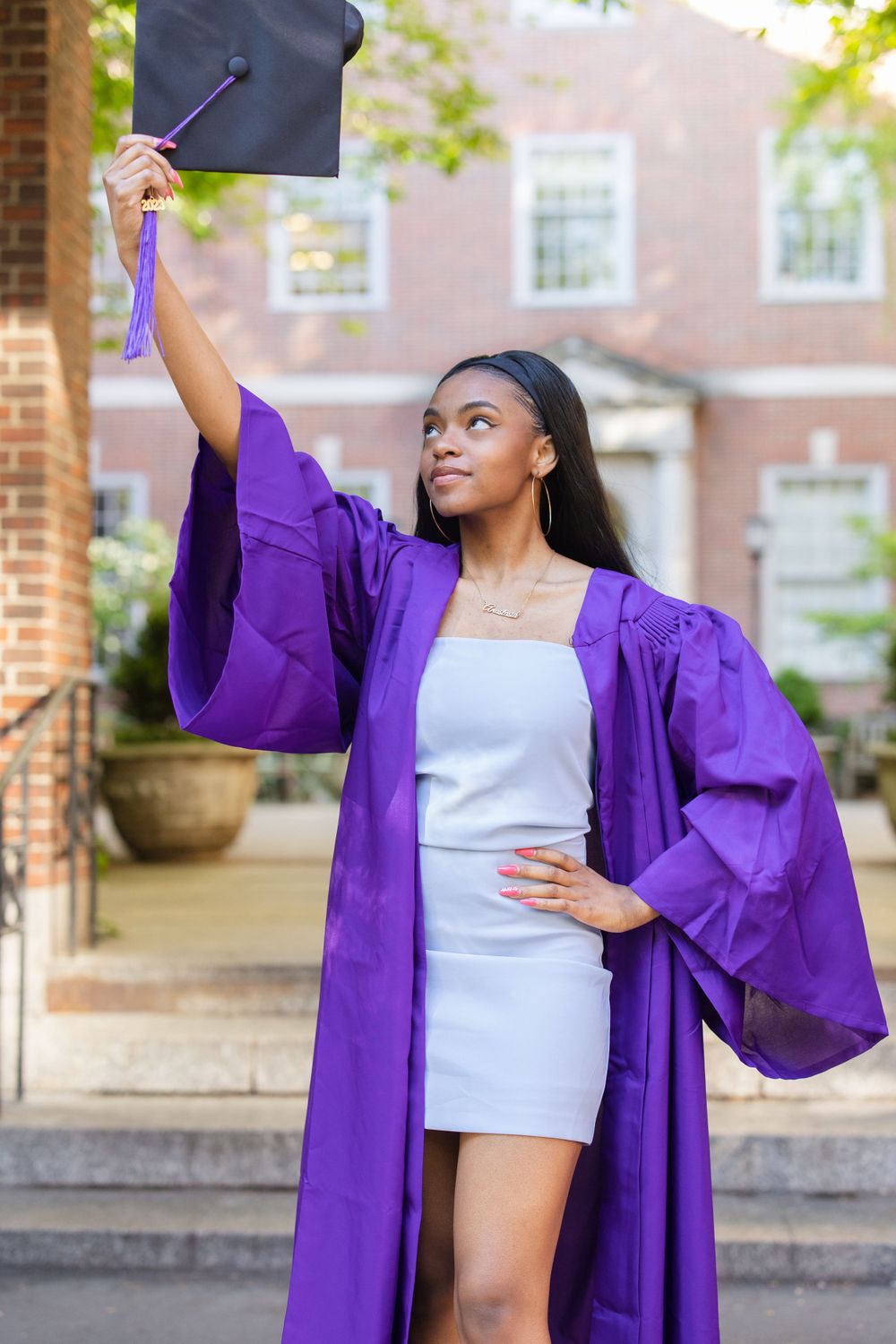 Graduate in purple regalia holds up graduation cap in celebratory pose.