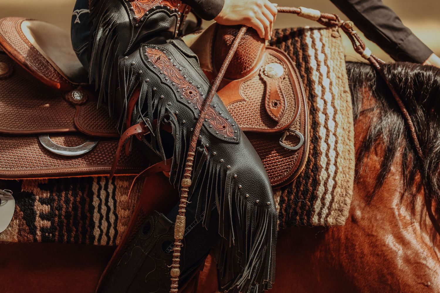 Close-up detail of brown leather western saddle with black fringe and decorative tooling and rope detailing.
