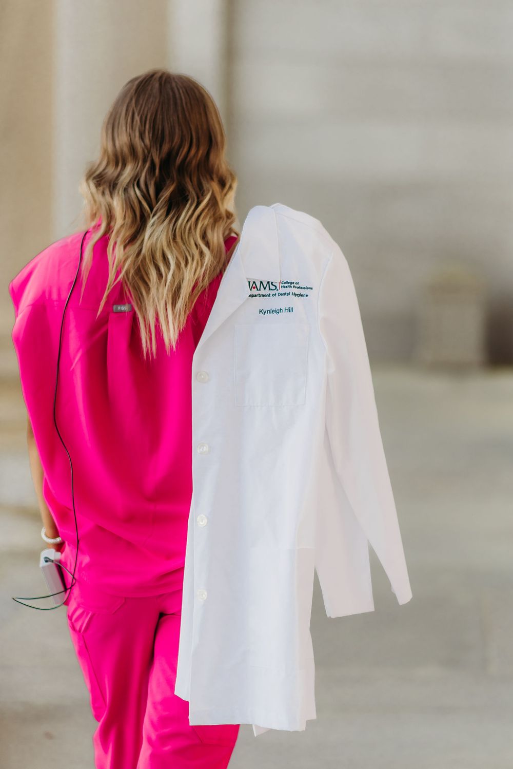 Back view of person in pink outfit holding white medical coat against neutral background.