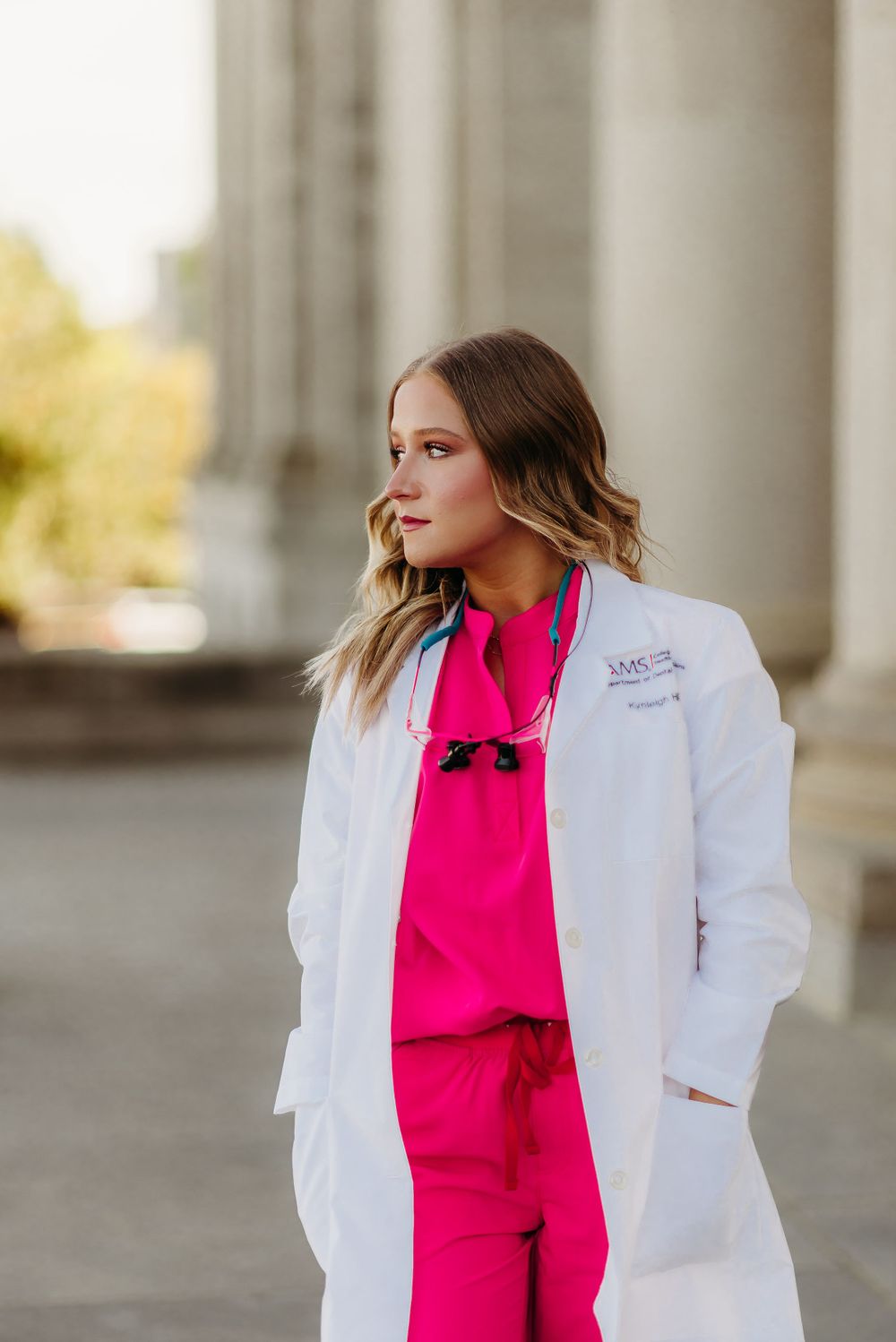 Person wearing white medical coat over bright pink scrubs stands in front of classical columns.