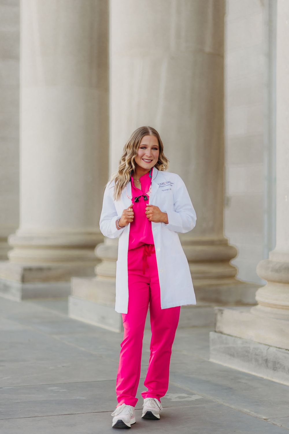 Medical professional in white coat and pink scrubs poses between classical stone pillars.
