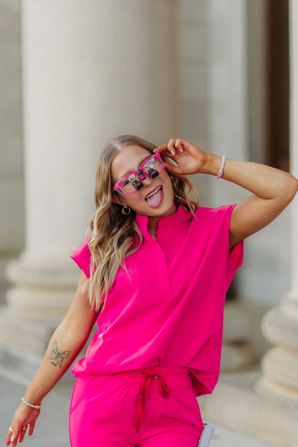 Energetic pose in hot pink scrubs and decorative sunglasses against architectural columns.