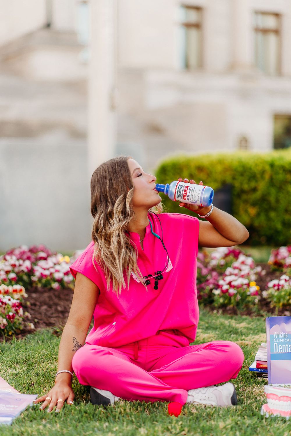 Person in pink scrubs drinks from water bottle while sitting on grass near flower bed.