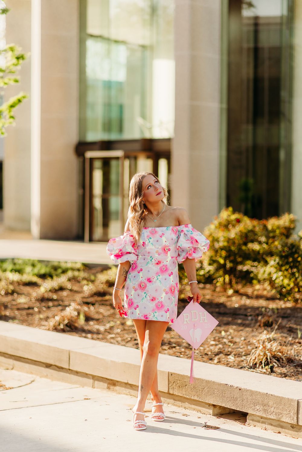 Person in floral off-shoulder dress stands confidently holding pink graduation cap outdoors.