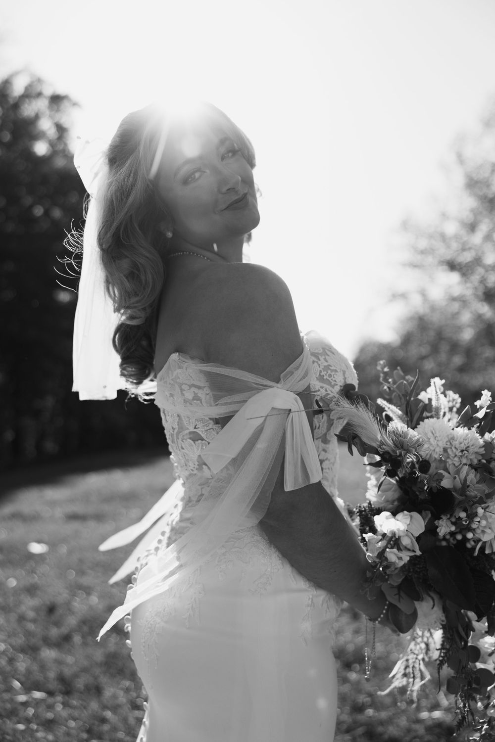 Elegant bride in lace wedding dress poses with bouquet in sunlit outdoor setting.