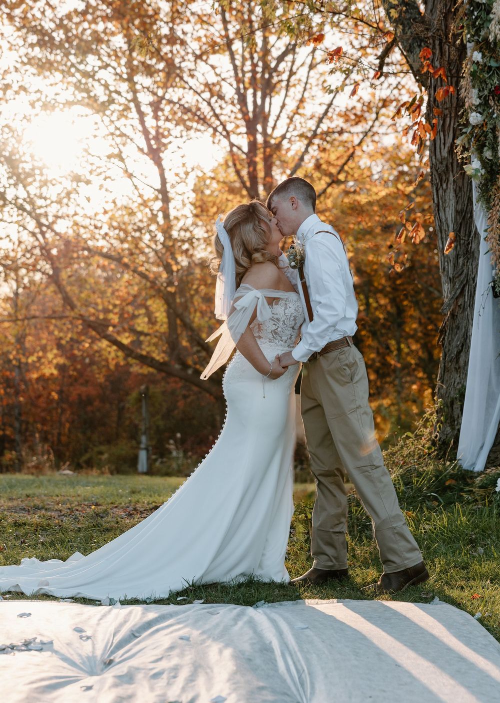 Bride and groom share a romantic kiss outdoors during golden hour, surrounded by vibrant autumn foliage.