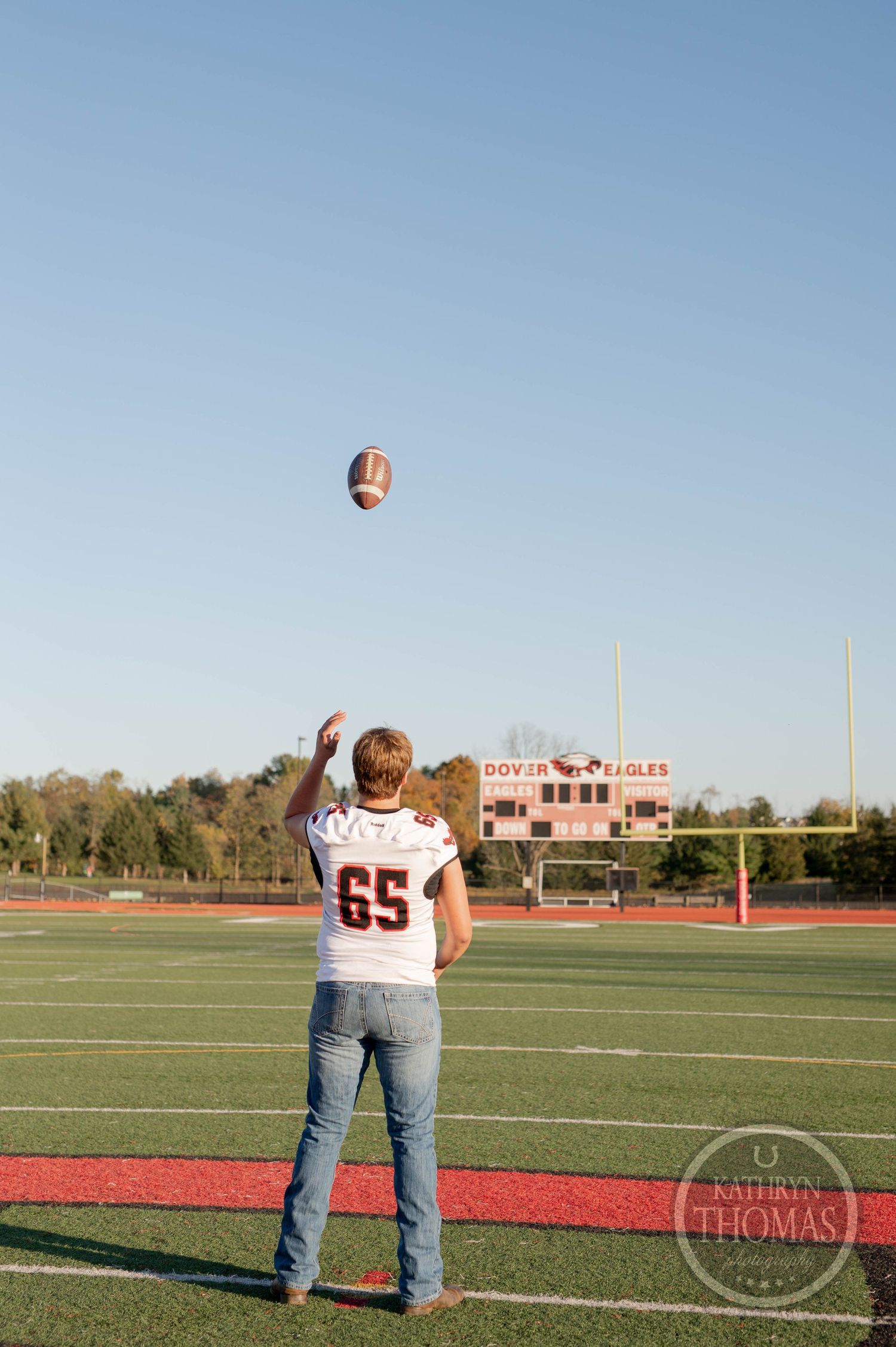 Player in jersey number 65 tosses football in air on sports field with stadium scoreboard visible.