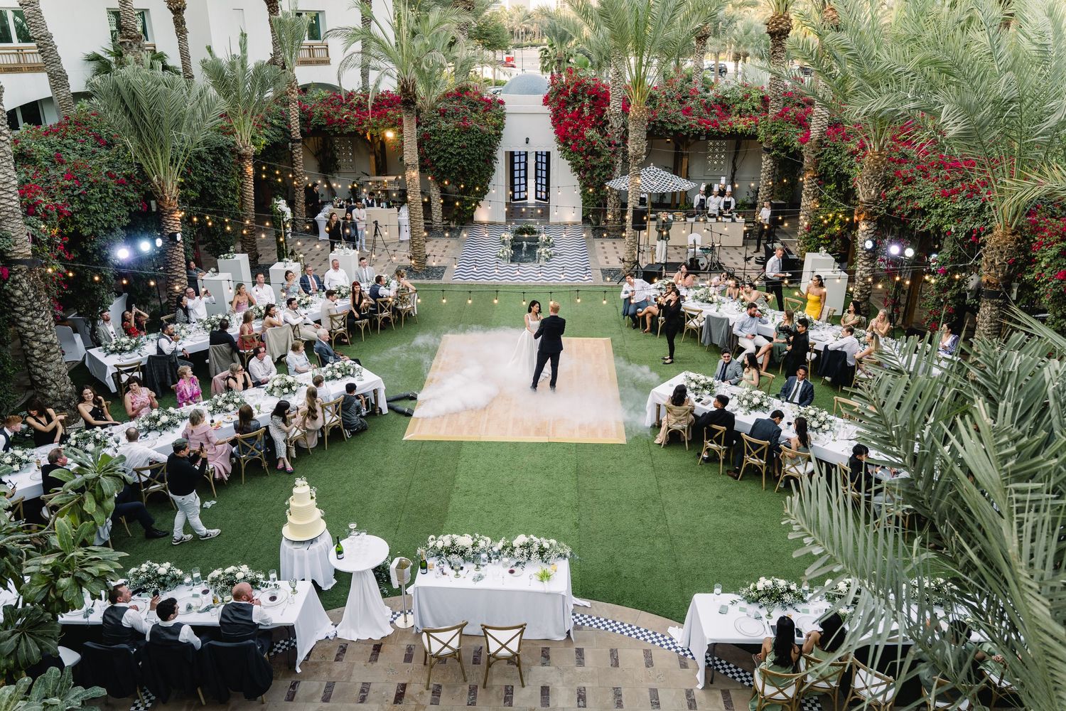 Aerial view of an elegant outdoor wedding reception with tables arranged around a green lawn dance floor surrounded by palm trees.