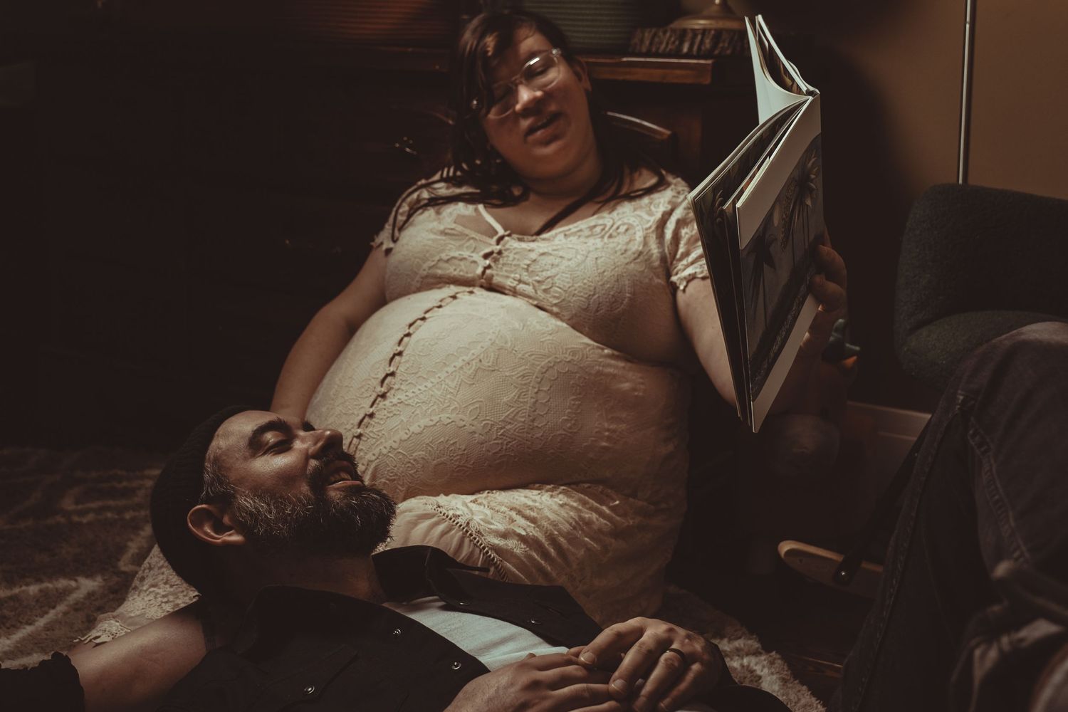 A couple sharing an intimate moment as one lies on a pregnant belly during a maternity photoshoot in dim lighting.