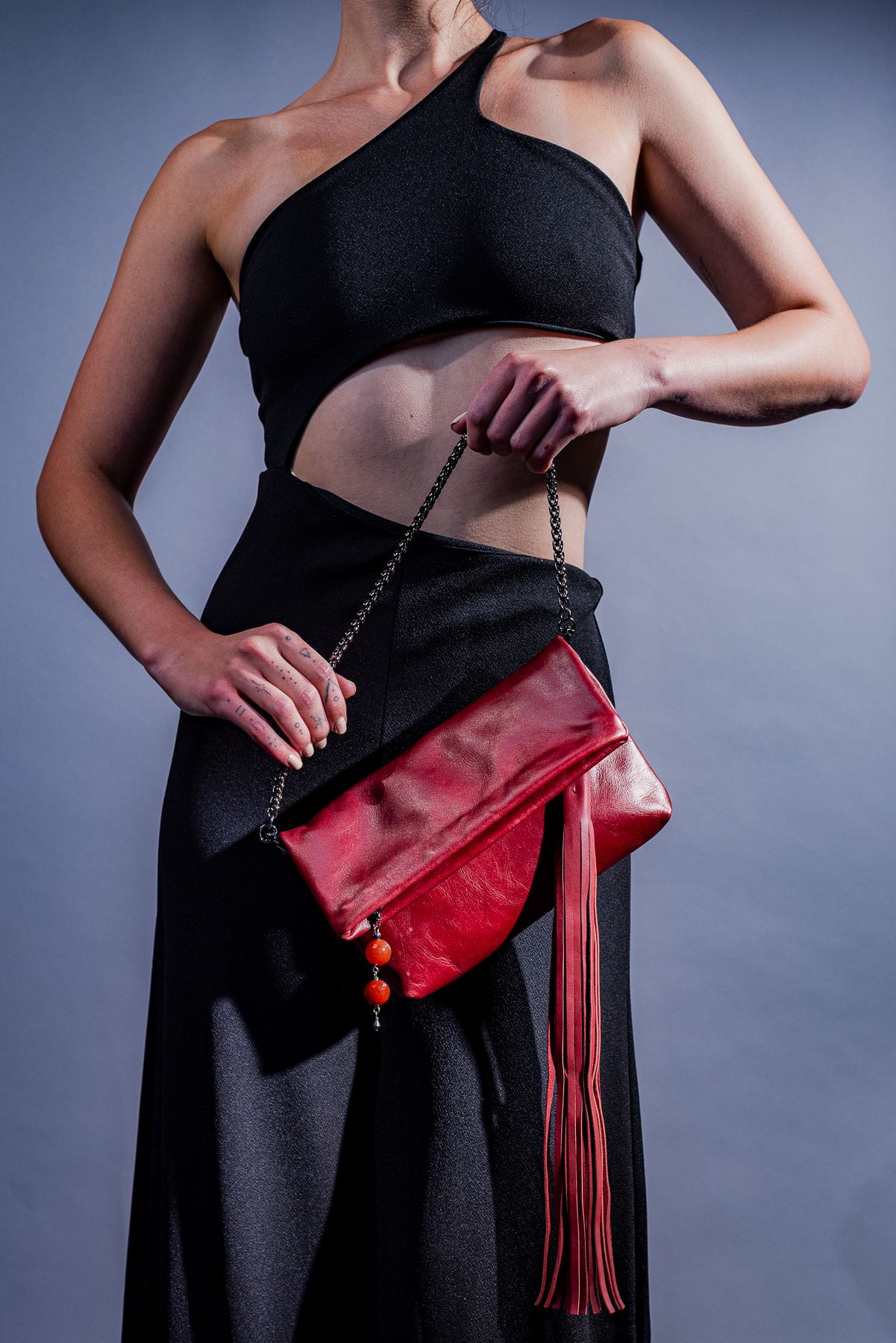 Person wearing black crop top and skirt holds red leather clutch bag with tassel detail against gray backdrop.