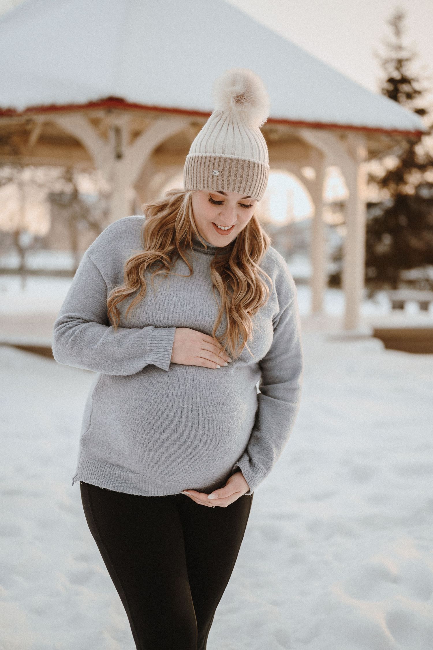 Expectant mother in winter wear smiles while touching her belly in snowy scenery.