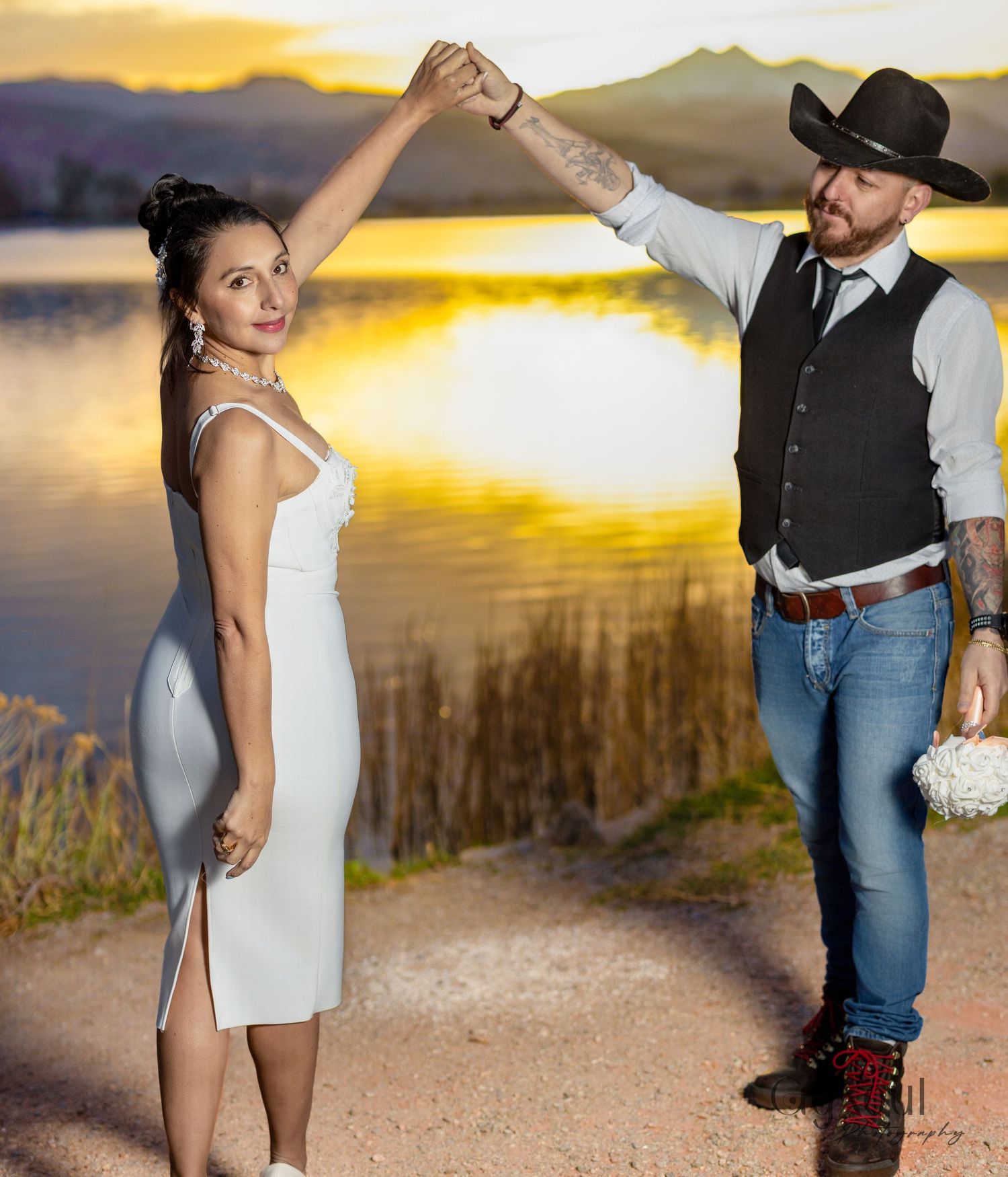 A couple in western attire holding hands against a golden sunset by a mountain lake.