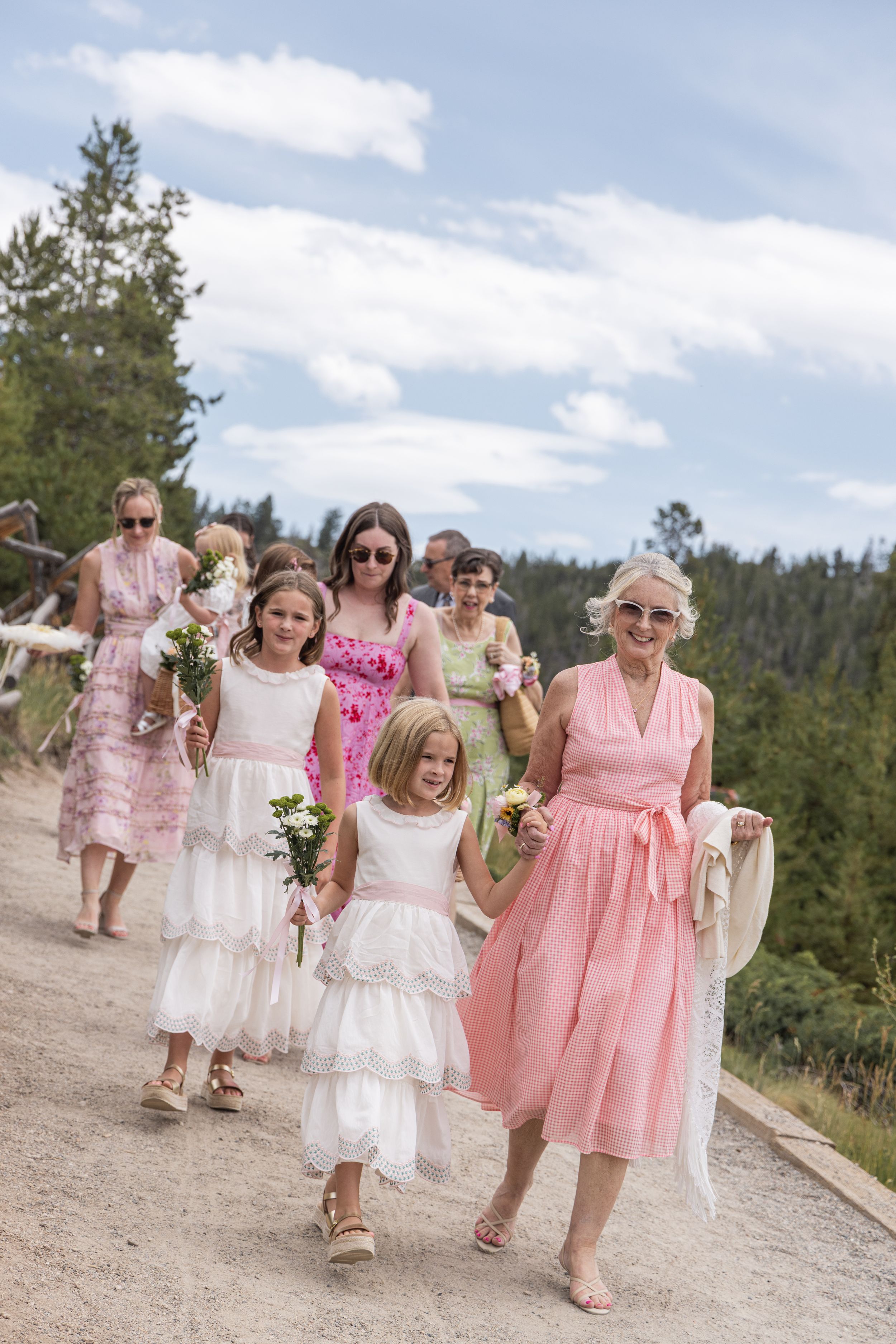 Group of guests in white and pink dresses walk along a dirt path during a sunny outdoor celebration.