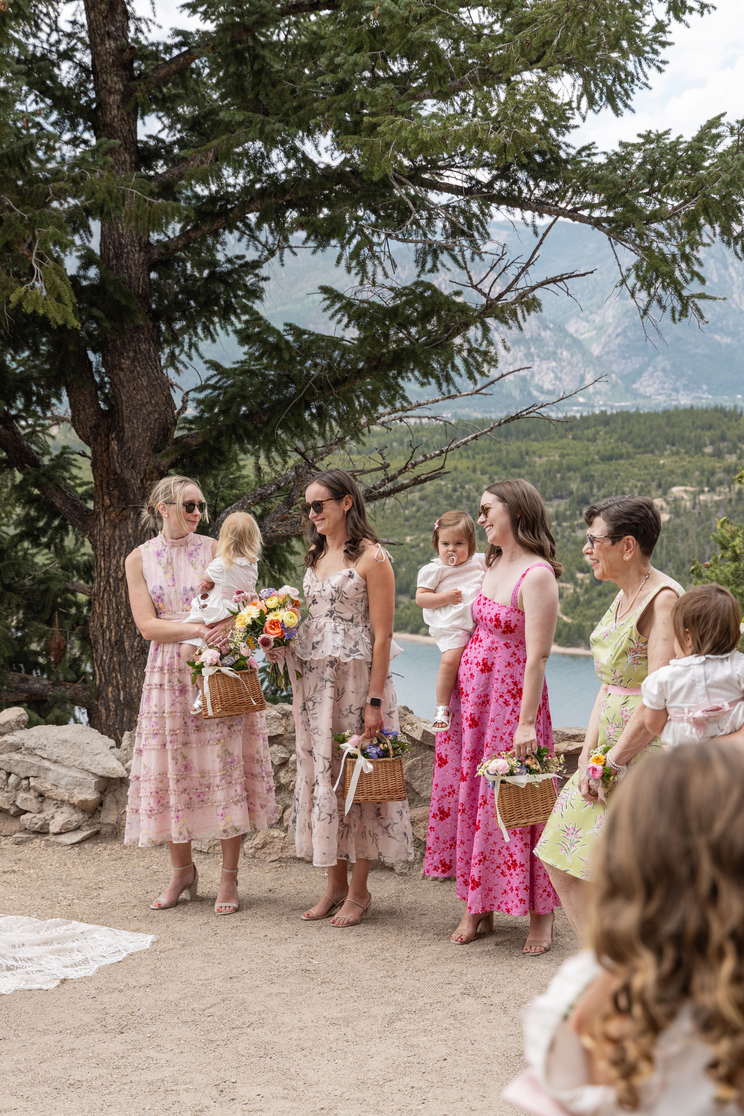 A group in floral dresses gathers at a scenic mountain overlook for a celebration on a sunny day.