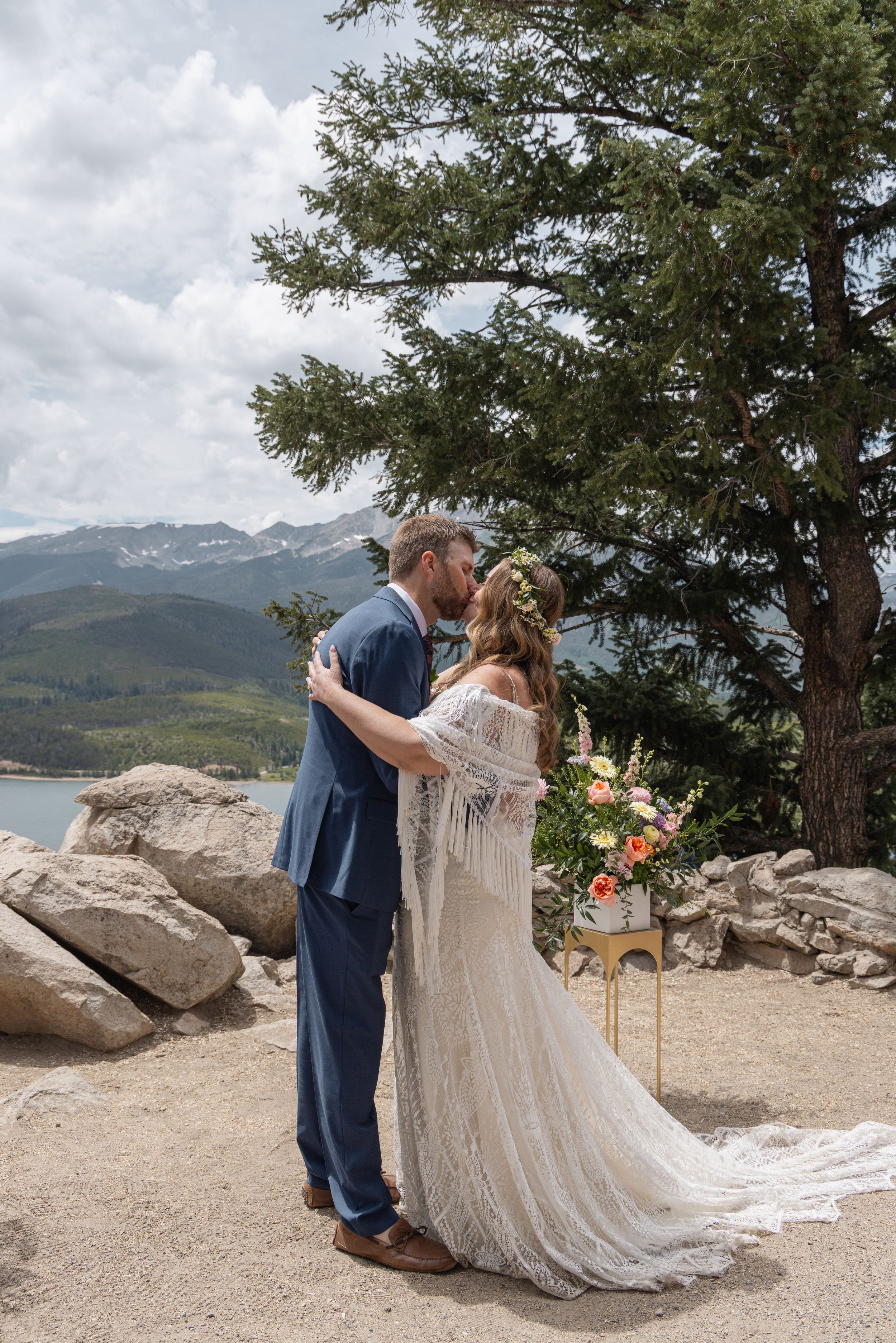 Couple sharing romantic moments on a mountain ledge overlooking a scenic lake and forested peaks during their wedding celebration.
