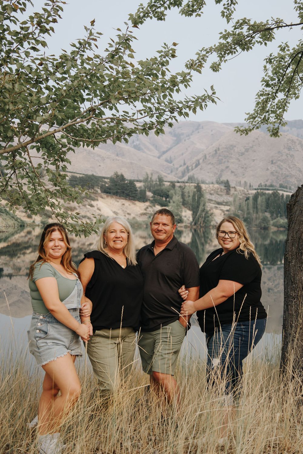 A group poses together against a scenic mountain lake backdrop in casual summer attire.