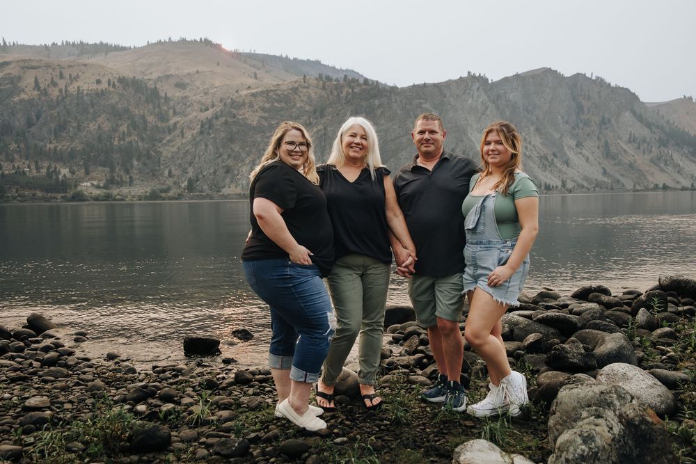 Four people posing together on rocky shoreline with mountains and lake in background.