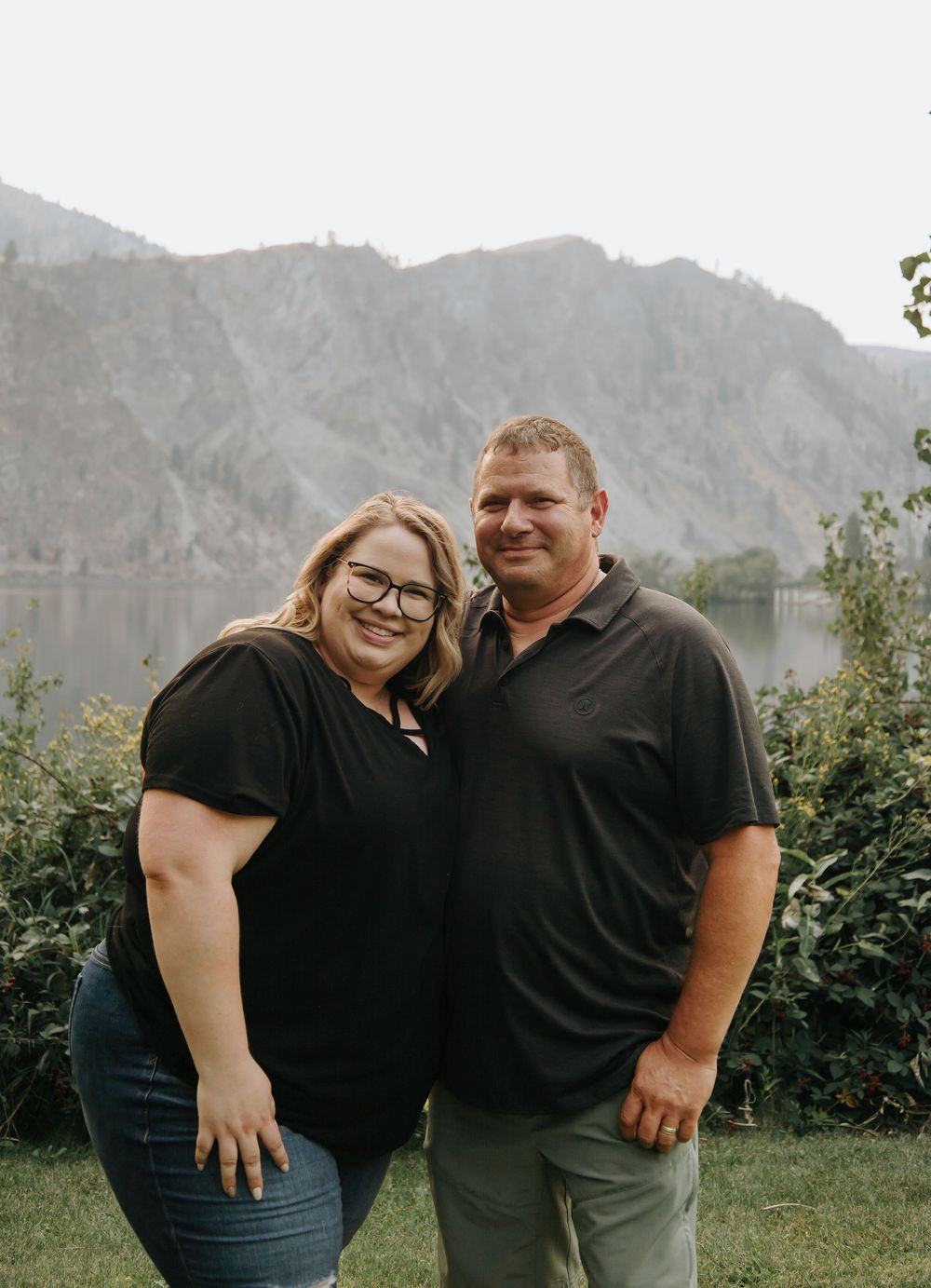 A couple poses together on grass with scenic mountain backdrop behind them.