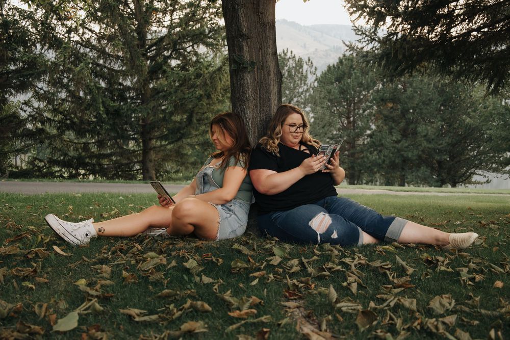 Two friends sitting back-to-back on grass during an outdoor photo session.