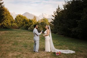 A couple in wedding attire holds hands in a grassy field with mountains and autumn trees in the background.