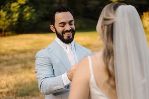 A groom in a light blue suit beams with joy during an outdoor wedding ceremony in a golden field at sunset.