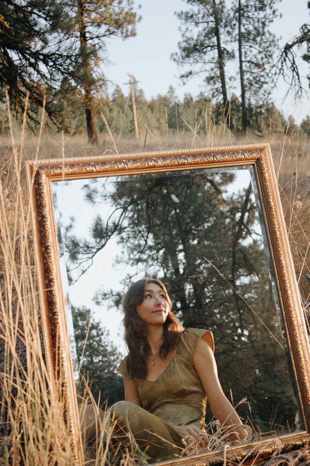 Artistic mirror portrait in a gold frame surrounded by tall grass and pine trees.