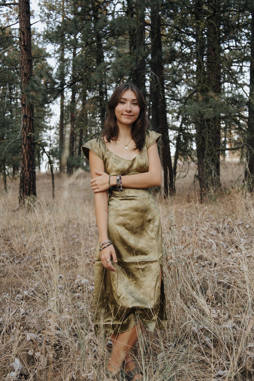 A woman in a gold dress poses in a forest clearing with tall pine trees in the background.