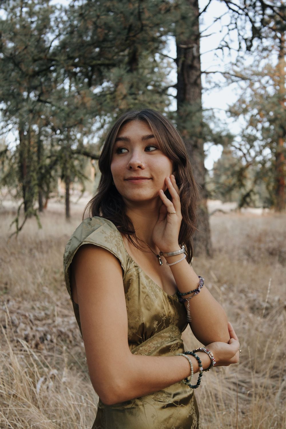 Portrait of a woman in olive dress standing among dry grass with pine trees behind.