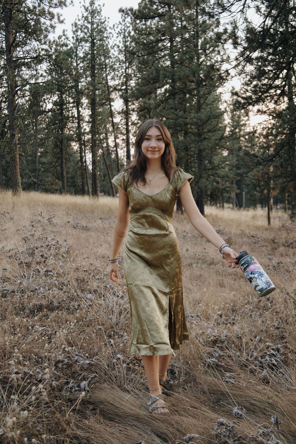 A woman walks through tall grass wearing a gold dress in a pine forest setting.