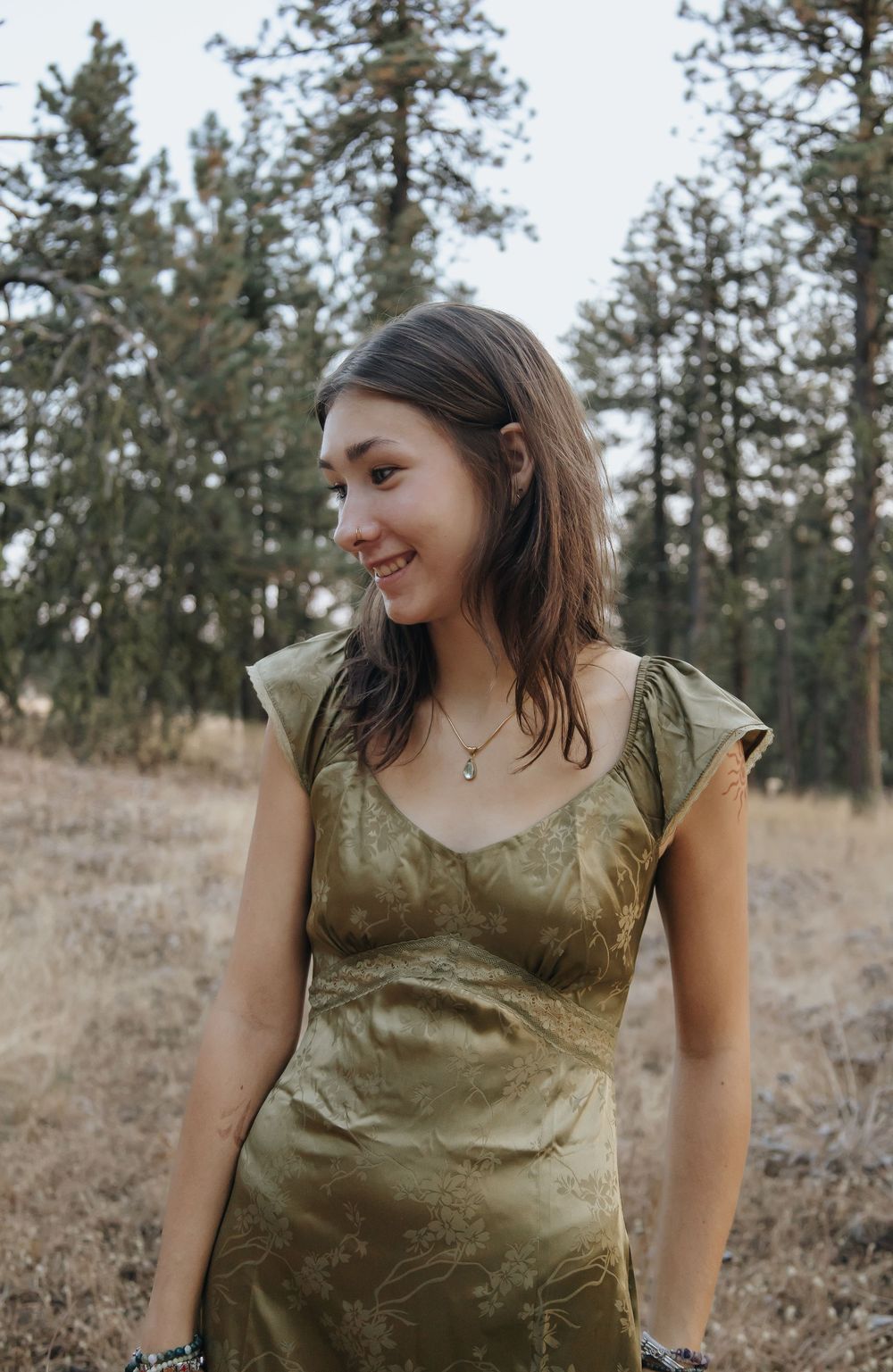 A candid shot of a woman in a sage green dress standing in a wooded area.