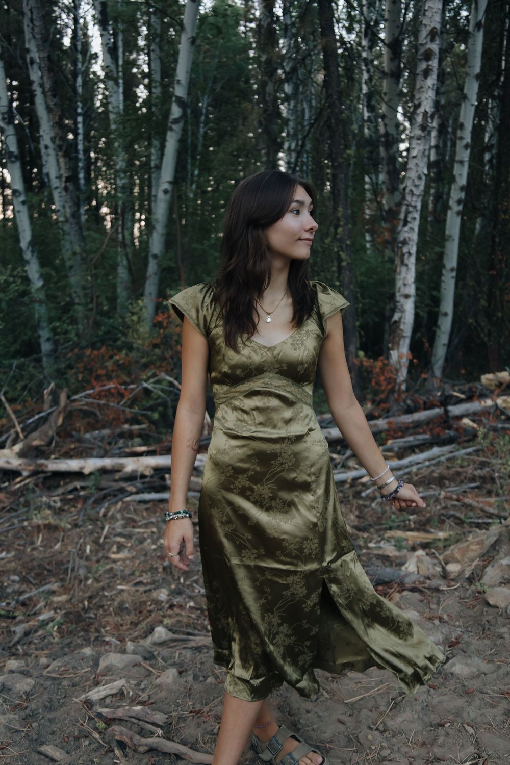 A woman in silk dress poses among birch trees in a moody forest setting.