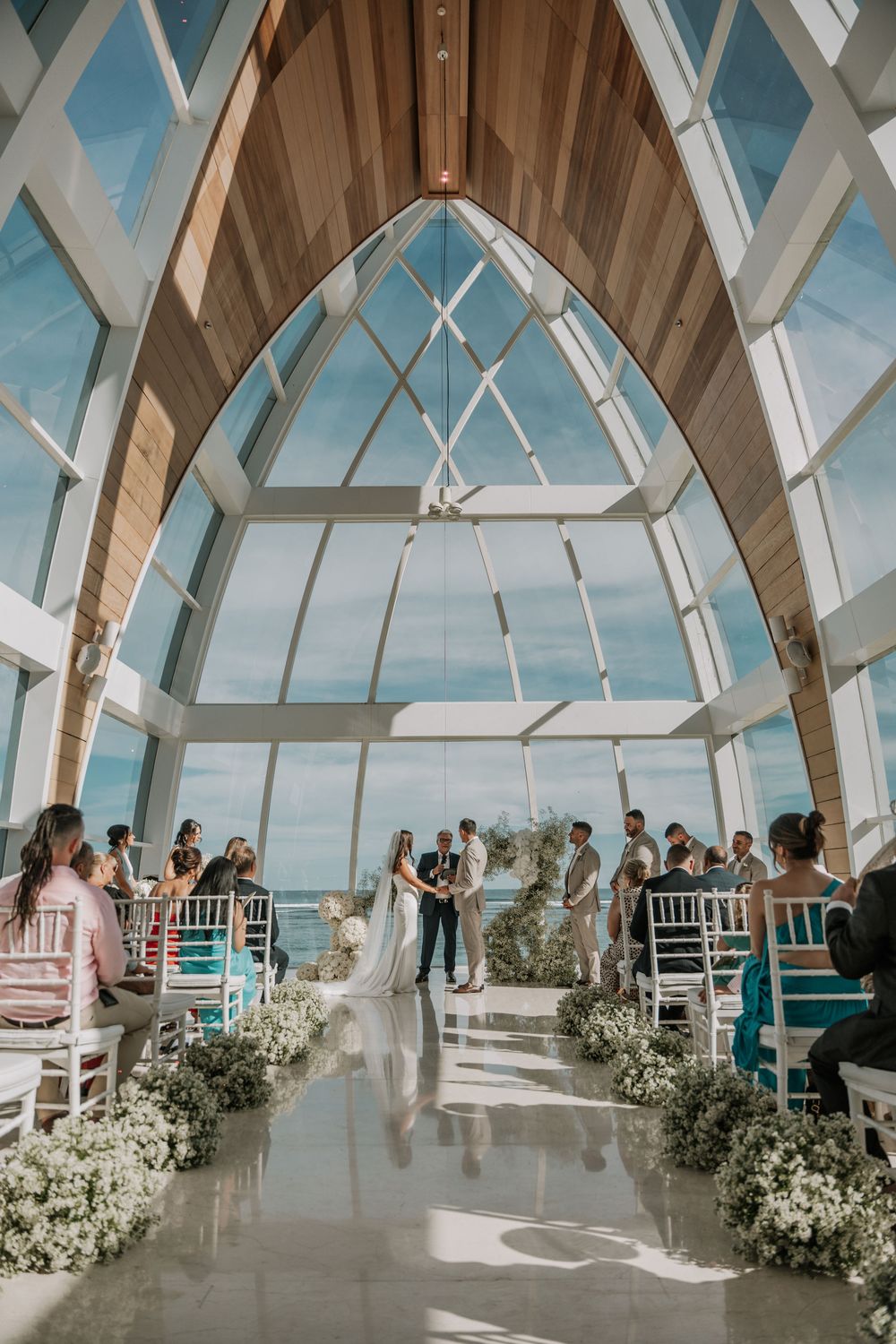 Wedding ceremony taking place in a modern glass chapel with geometric windows and white floral decorations.