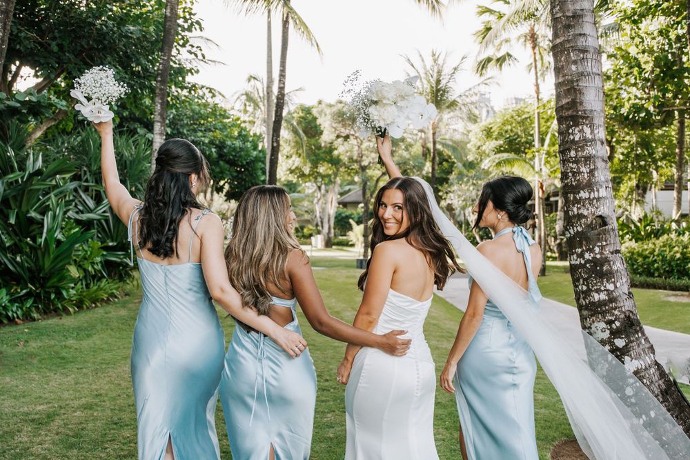 Bridesmaids in light blue dresses embrace and laugh together in a tropical garden setting with palm trees.