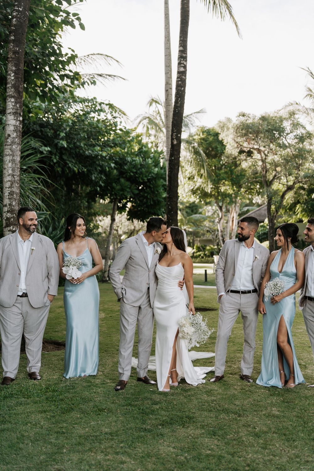 Wedding party in light gray suits and powder blue dresses walk in line on green lawn under palm trees at sunset.