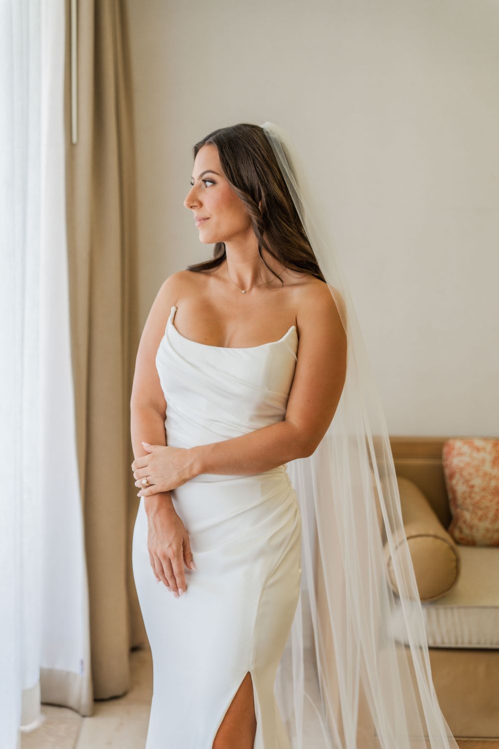Bride wrapped in white fabric and veil gazes elegantly out window in natural light bridal portrait session.