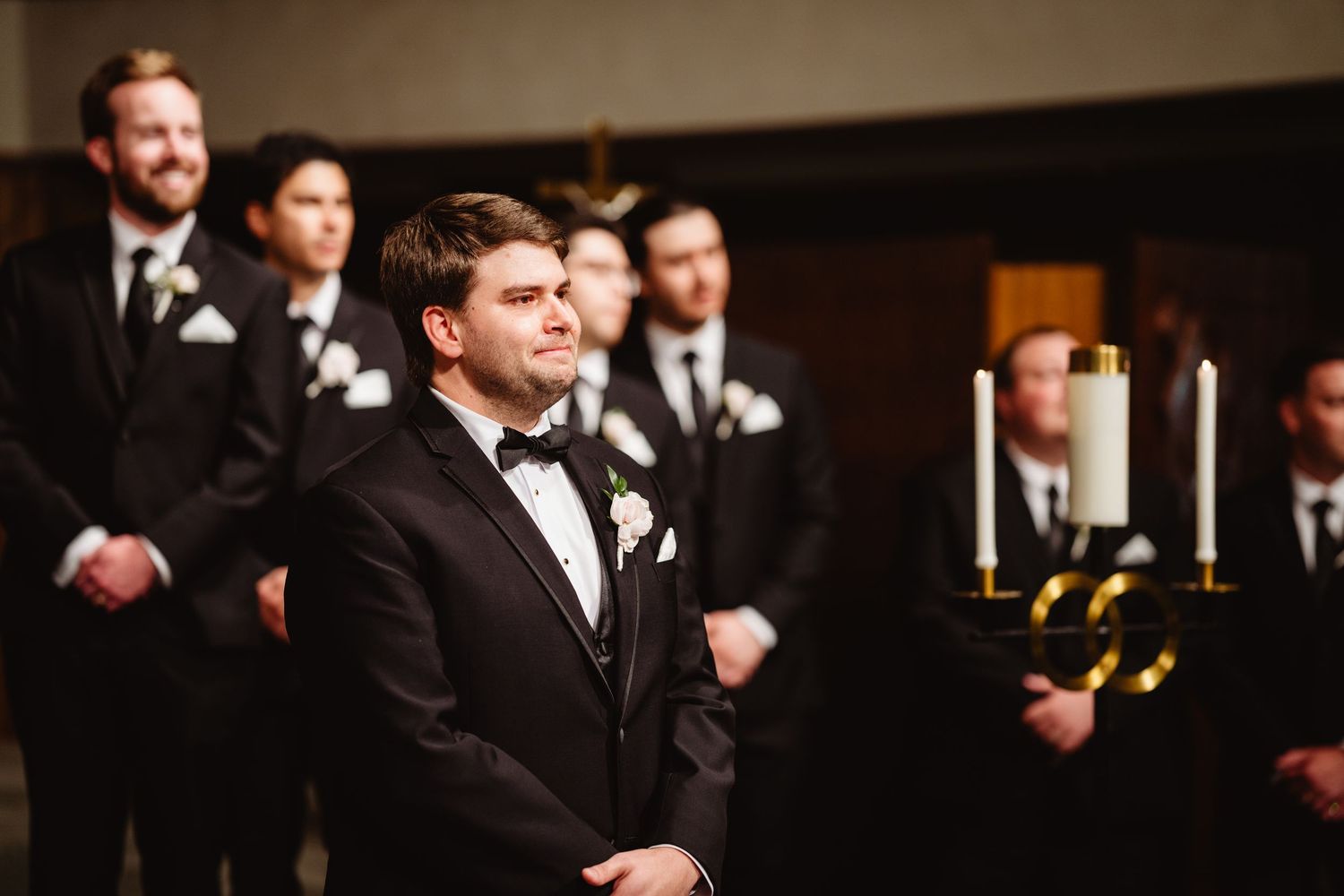 Wedding attendees in formal black suits and boutonnieres smiling during a joyful celebration at an indoor venue.