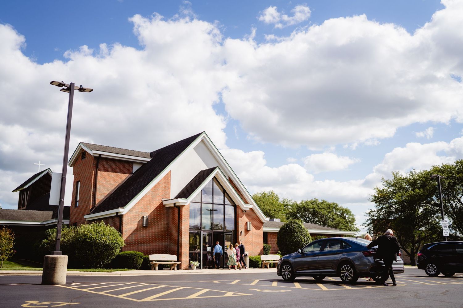 Modern brick church buildings with white accents and angled rooflines stand against a blue sky with puffy white clouds.