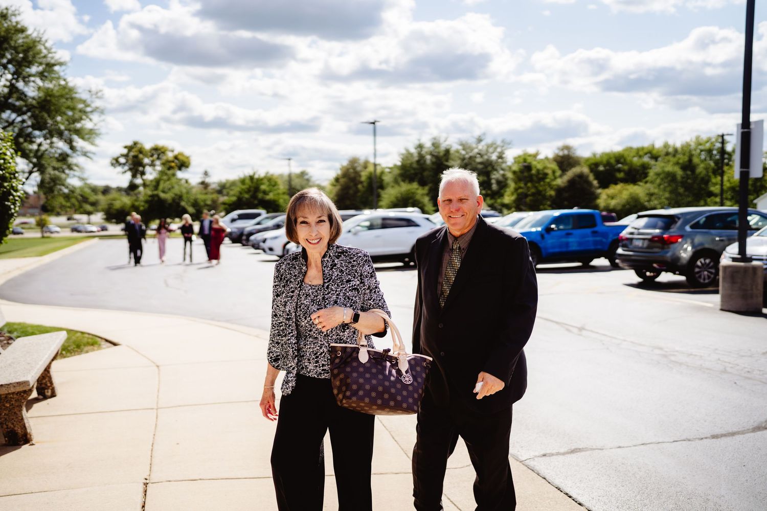 Two people walk together along a paved walkway on a sunny day with cars parked in the background.