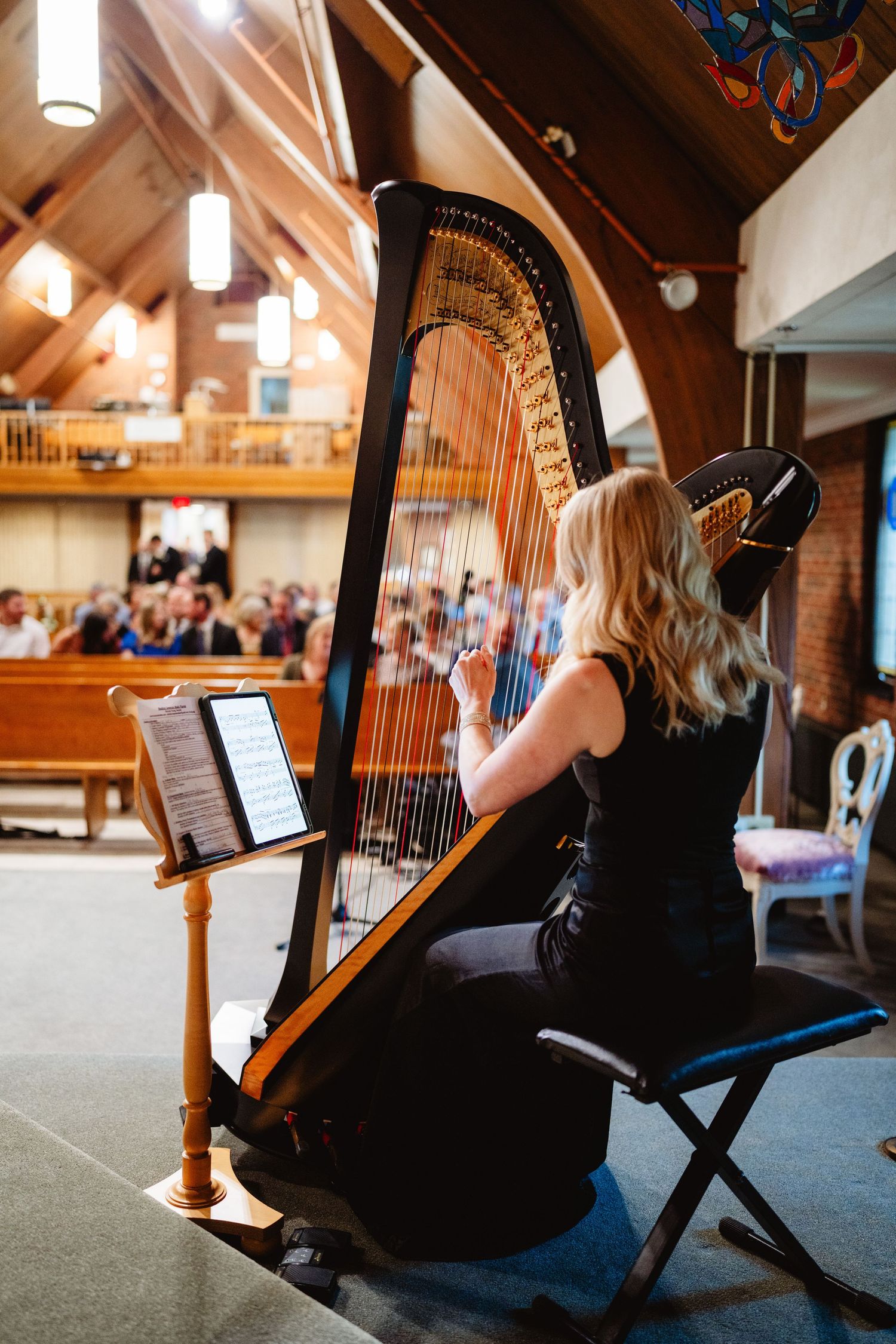 A harpist in a black dress performs in a wooden church hall with warm lighting and sloped ceiling.