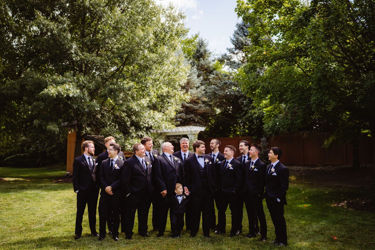 Wedding party stands outdoors in dark suits while lined up against a natural backdrop with trees and greenery.